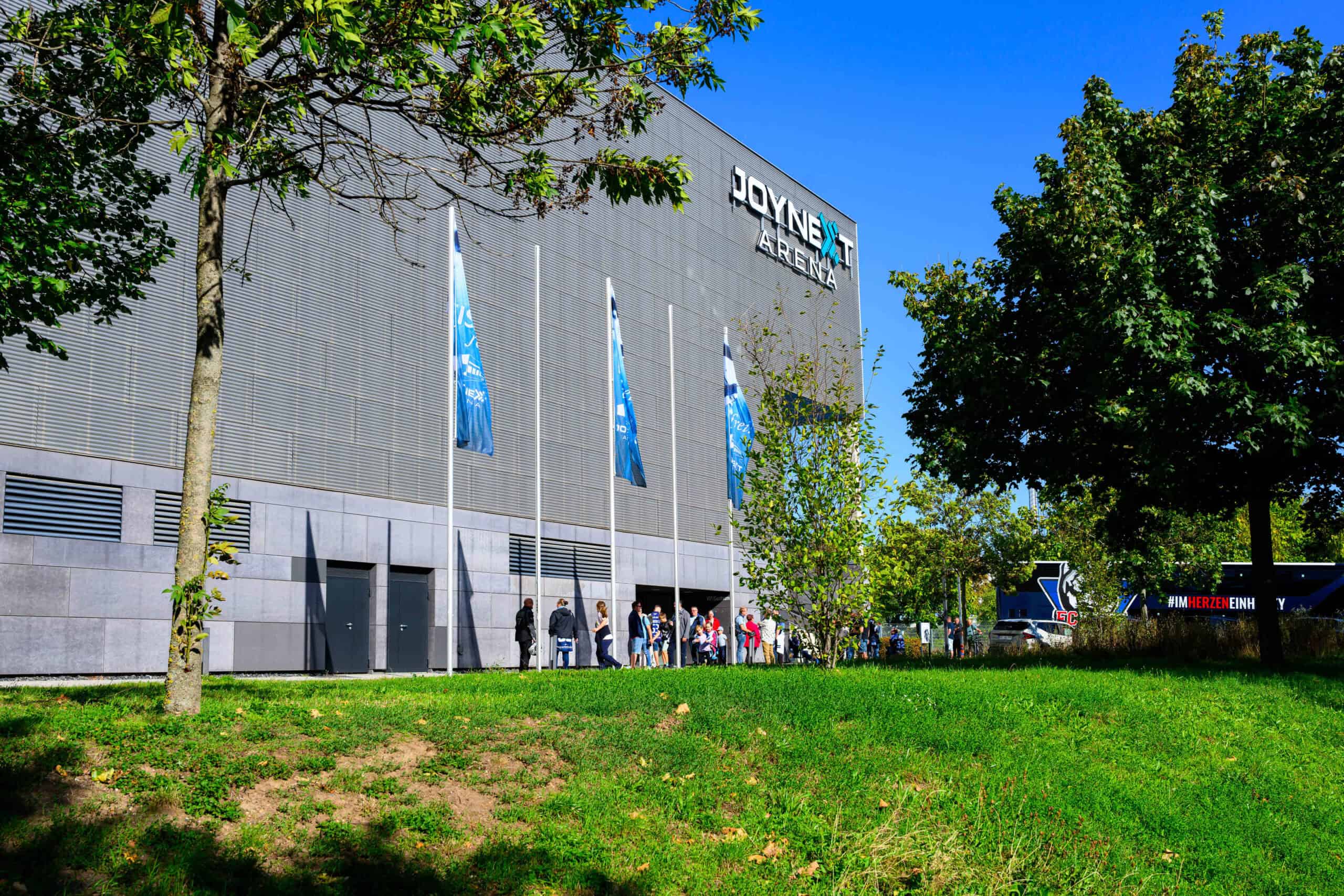 Die JOYNEXT Arena von außen an einem Sommertag, davor Bäume und Fahnen